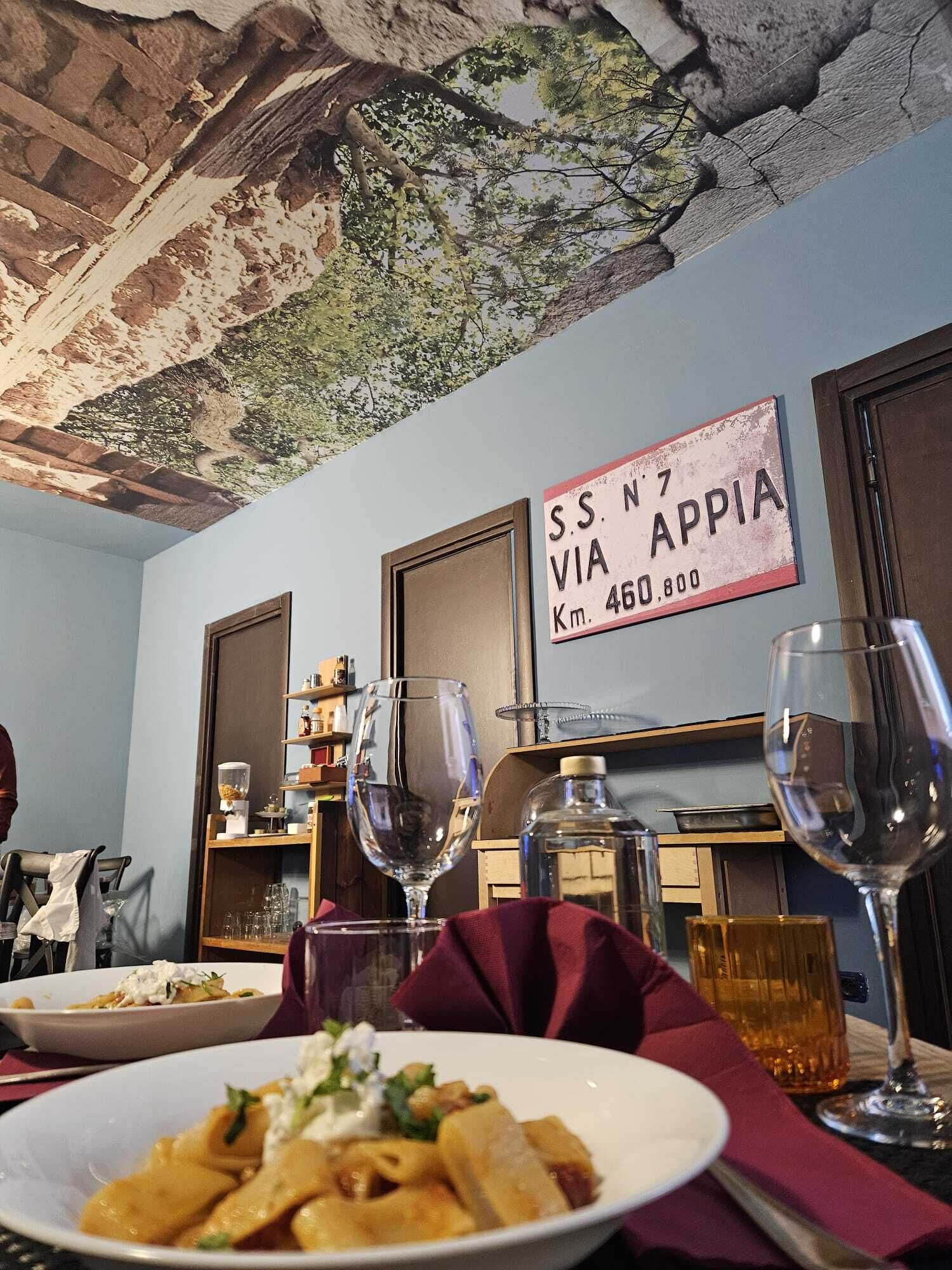 A dining area with a rustic ceiling, a sign indicating 'SS N7 VIA APPIA Km. 460,800', and a table with plates of food and wine glasses.