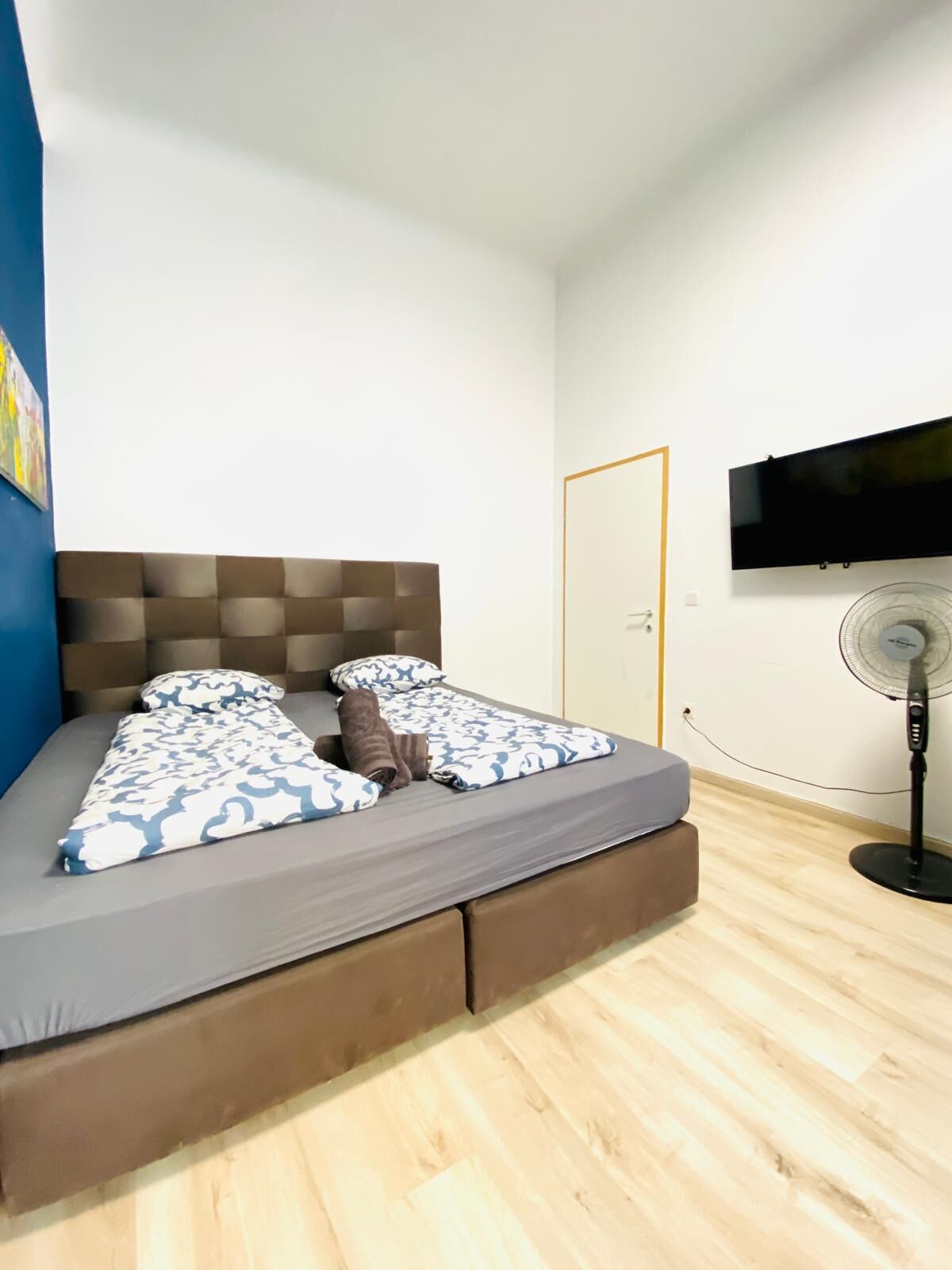 Una cama individual con cabecera, una mesita de noche, un televisor instalado en la pared y un ventilador de pie.