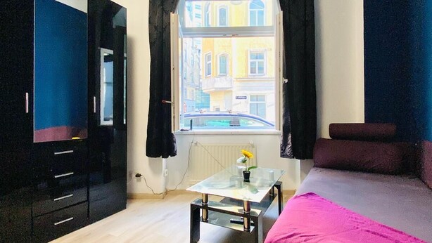 Ein Zimmer mit einem Bett, einem Tisch mit einer Vase, einem Fernseher und einem Fenster mit Vorhängen.