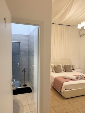 Una habitación de hotel moderna con una cama grande, una mesita de noche y un tocador. Cuenta con un baño con ducha y puerta de vidrio.