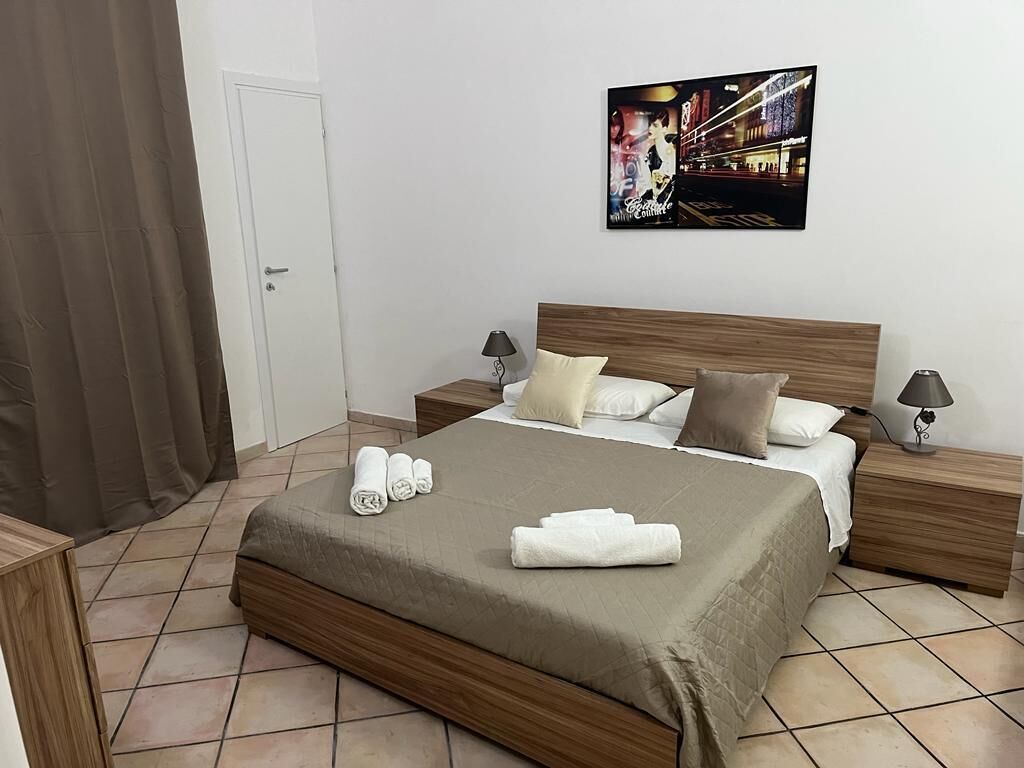 Cama bem arrumada com cabeceira marrom, duas mesas de cabeceira com abajures e um quadro na parede.