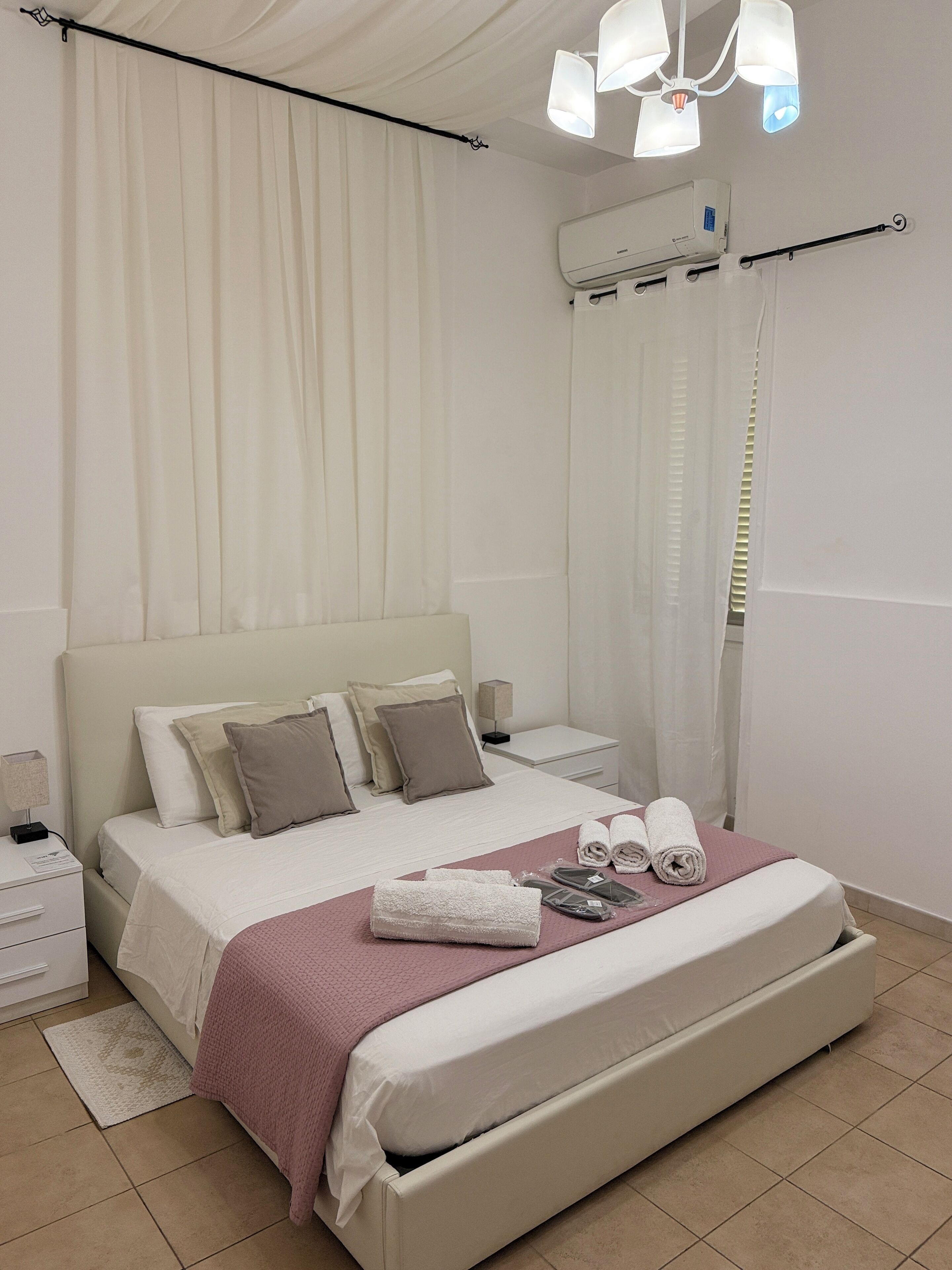 A neatly arranged bedroom with a bed, pillows, a bedside table, a lamp, and a window with curtains.