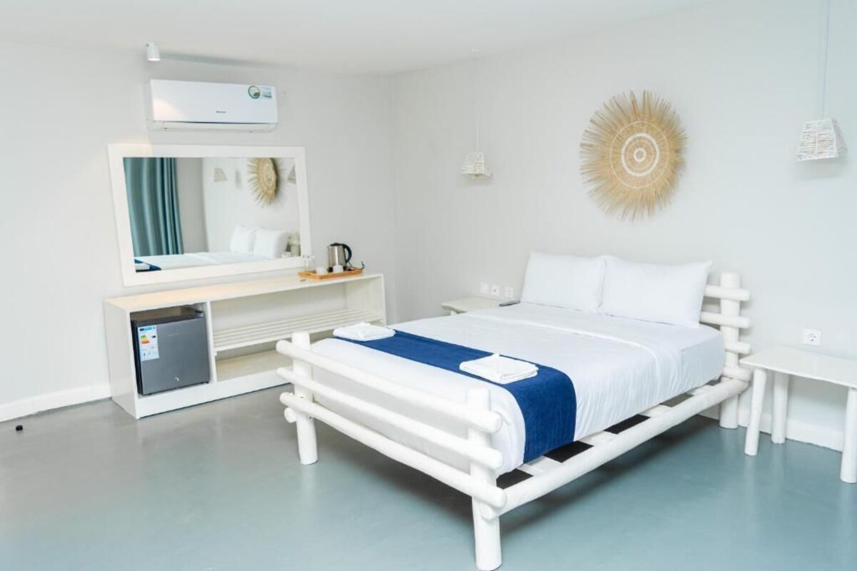 A modern hotel room with a white bed, a small bedside table, a built-in mini-fridge, a large mirror, and a decorative wall piece.