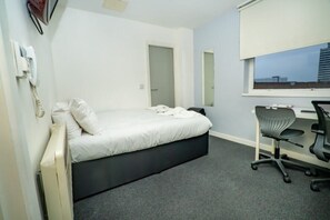 Ein Einzelbett mit weißer Matratze und dunklem Kopfteil, ein Schreibtisch mit Stuhl, ein Fenster mit Blick und ein fest montiertes Telefon an der Wand.