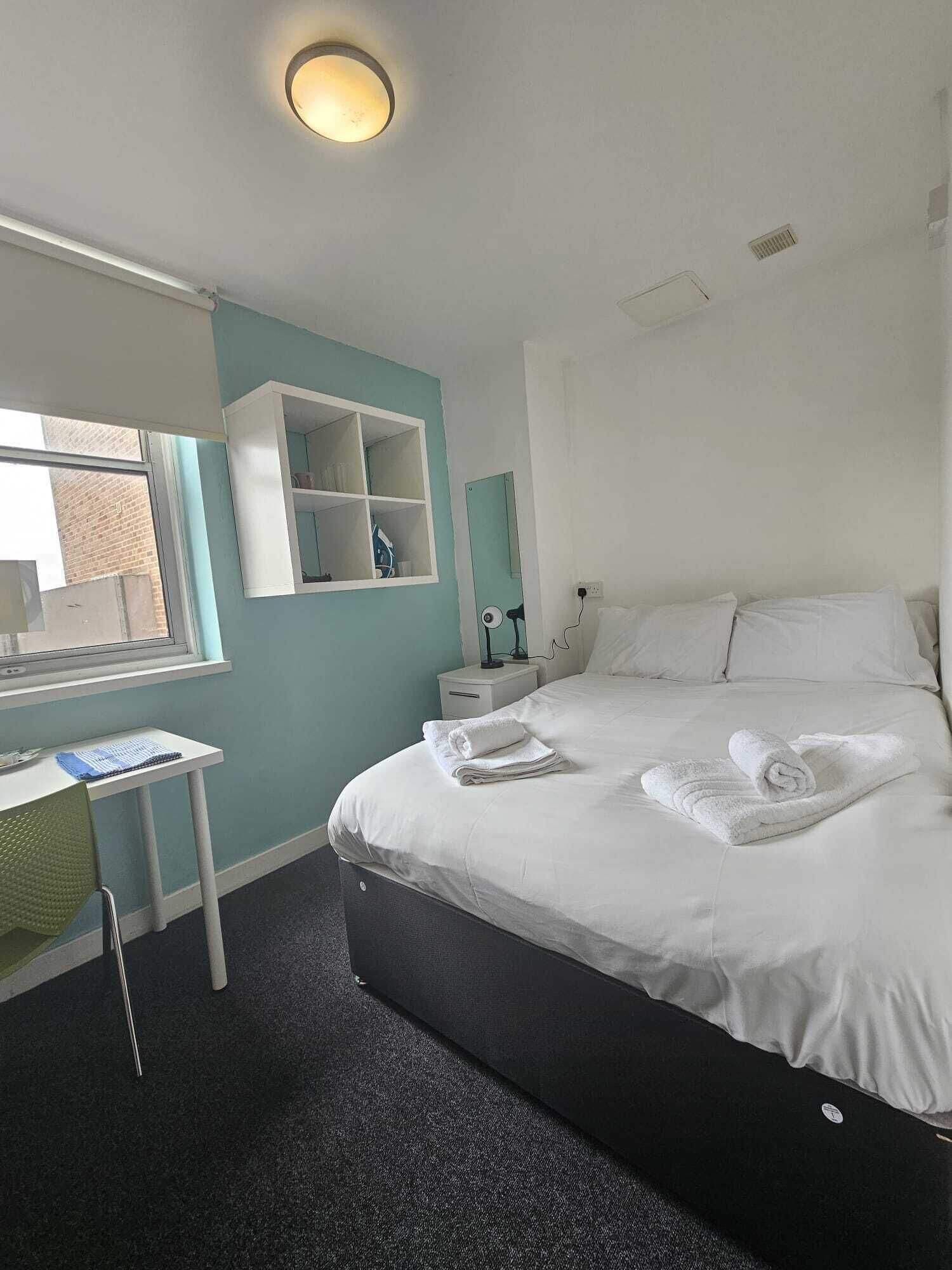 A hotel room with a bed, a desk, a chair, a window, and a shelf.