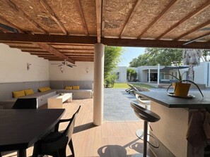 Un espace extérieur aménagé en terrasse, avec un plafond en bois, un coin salon doté de coussins, une table à manger et un comptoir de bar.