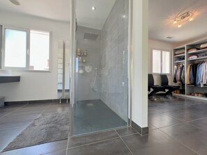 Une salle de bain moderne dotée d’une douche à parois de verre, d’un dressing avec des étagères bien organisées et d’une grande fenêtre laissant entrer la lumière naturelle.