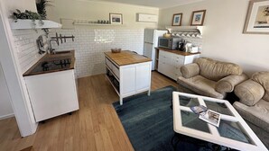 Kompakti keittiö, jossa on valkoiset kaapit, puinen työtaso ja sisäänrakennettu mikroaaltouuni. Olohuoneessa on beige sohva ja lasinen sohvapöytä.