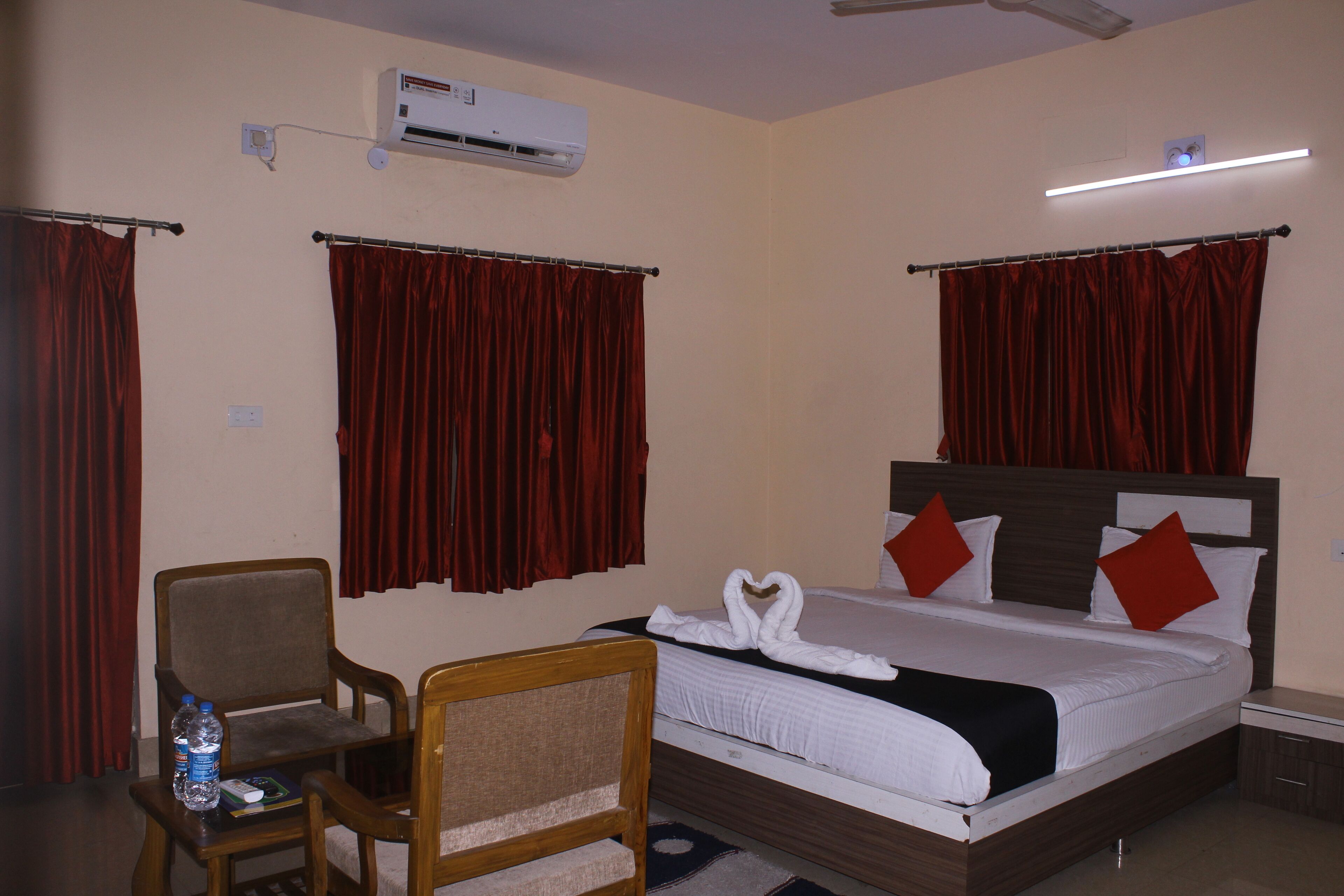A hotel room with a bed, two chairs, a table with a water bottle, an air conditioner, and a ceiling fan.