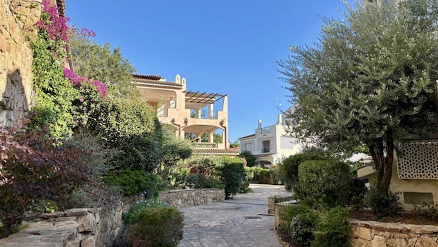 Un estrecho sendero de piedra bordeado de abundante vegetación y edificios de estilo mediterráneo tradicional.