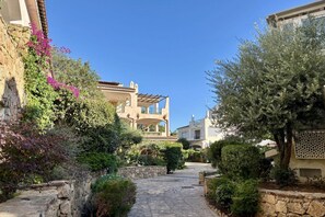 A narrow stone pathway flanked by lush greenery and traditional Mediterranean-style buildings. - Porto Rotondo Studio 3 switching possibilities (Porto Rotondo)
