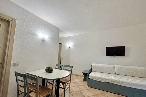 A small, clean room with a dining table, chairs, a sofa, and a TV mounted on the wall. - Porto Rotondo Studio 3 switching possibilities (Porto Rotondo)