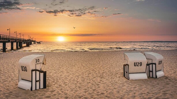 Una playa con dos sillas de playa identificadas con las etiquetas U30 y U29, un muelle al fondo, y el sol poniéndose sobre el océano.