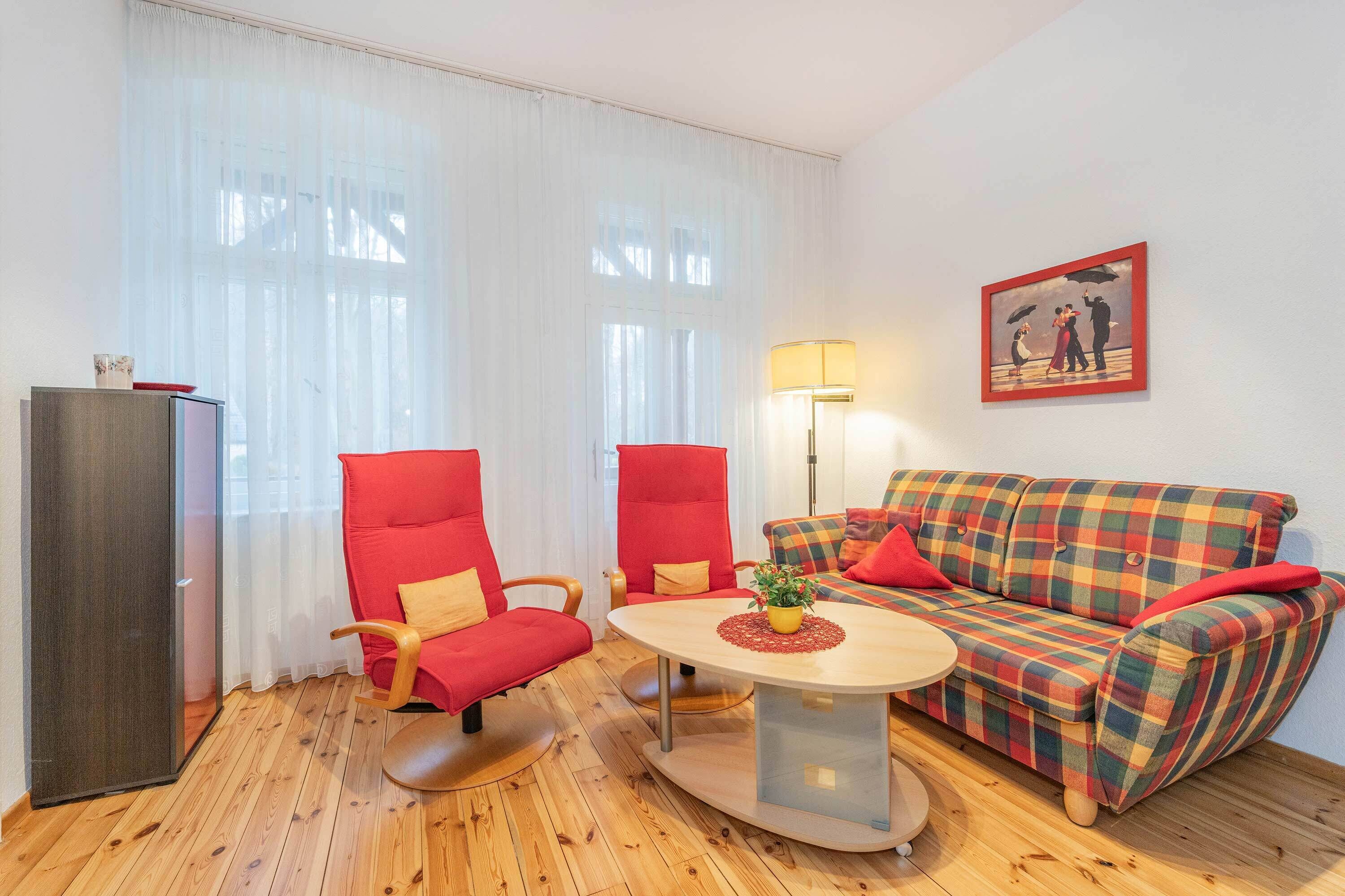Una habitación con piso de madera, un sofá rojo, una silla roja, una pequeña mesa, una lámpara y un cuadro en la pared.