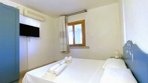 Hotellihuone, jossa on sänky, seinälle asennettu televisio, ilmastointilaite, ikkuna verhoilla sekä pieni pöytä lampun kanssa.