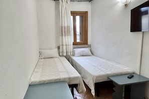 Une petite chambre compacte avec deux lits simples, une fenêtre avec un rideau, un téléviseur à écran plat fixé au mur et une petite table avec une télécommande.