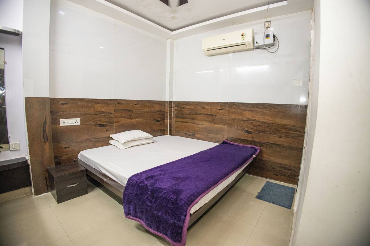A bedroom with a bed, a wooden headboard, a white wall, and an air conditioning unit.