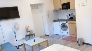 Ein kompakter Wohnbereich mit Sofa, kleinem Tisch, Ventilator, Waschmaschine und Mikrowelle im Küchenbereich.
