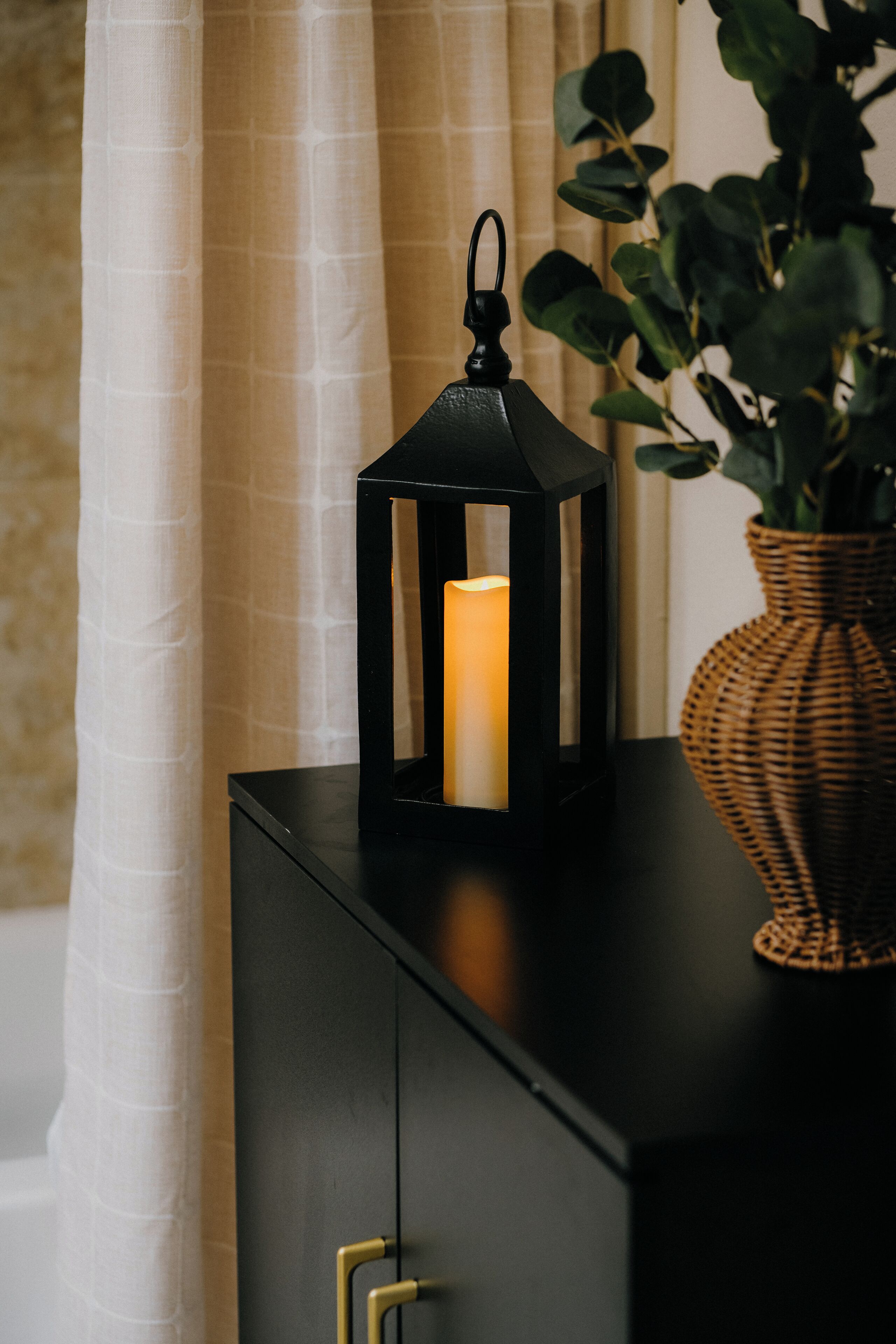 Un mueble negro con una vela dentro de un farolillo, un florero de mimbre con hojas verdes y una cortina blanca al fondo.