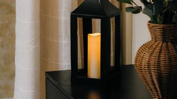 Un mueble negro con una vela dentro de un farolillo, un florero de mimbre con hojas verdes y una cortina blanca al fondo.