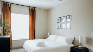 A neatly made bed with white linens, a bedside table with a lamp, and a wall with framed pictures. - Downtown Loft • Balcony • King Bed • Stylish Stay (Motgomery)