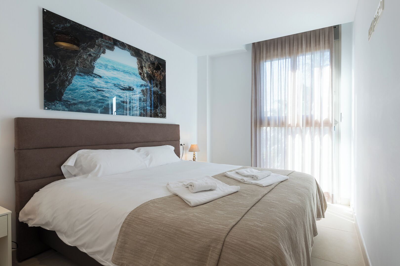 Un letto rifatto con lenzuola bianche e beige, un grande quadro raffigurante una scena costiera sopra, e una finestra con tende leggere che lasciano filtrare la luce naturale.