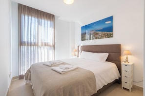 Ein ordentlich bezogenes Bett mit Kopfteil, ein Nachttisch mit Lampe und ein Fenster mit Vorhängen.