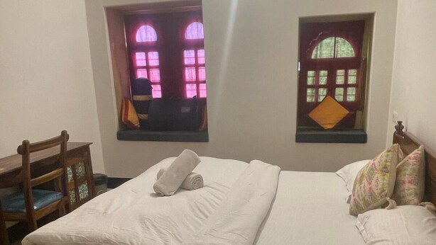A neatly made bed with a rolled towel, two windows with curtains, a wooden chair, and a patterned rug. - The Ginger House (Jaisalmer)