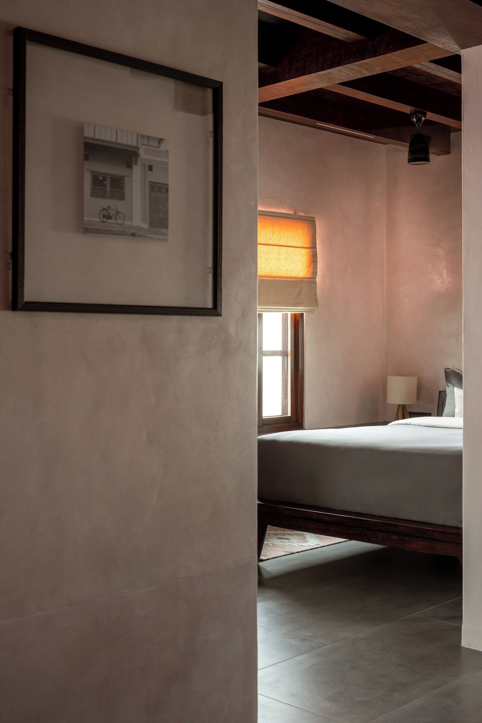 A hotel room with a bed, a framed picture on the wall, a window with a view of a building, and a lamp on the nightstand.