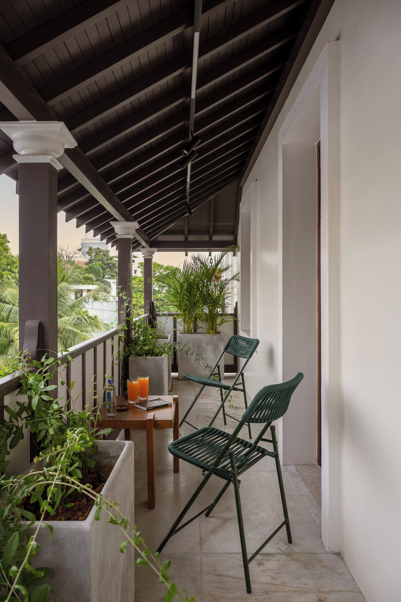 A covered balcony with a table and chairs, potted plants, and a view of greenery.