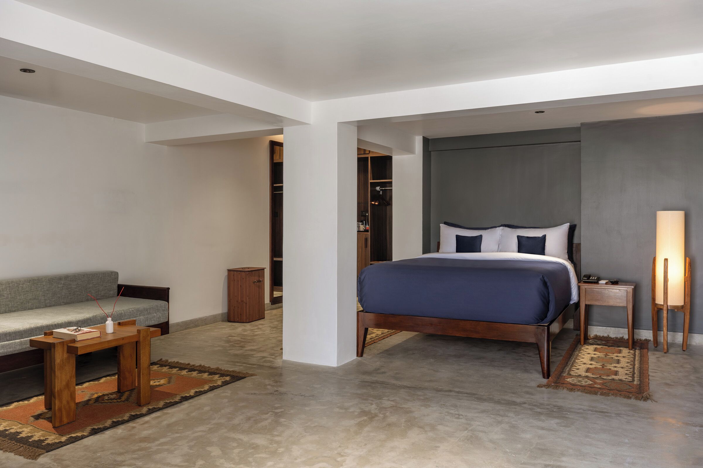 Une chambre d’hôtel moderne avec un grand lit, une table de chevet en bois, une lampe de chevet, un canapé et une petite table sur laquelle est posé un vase.
