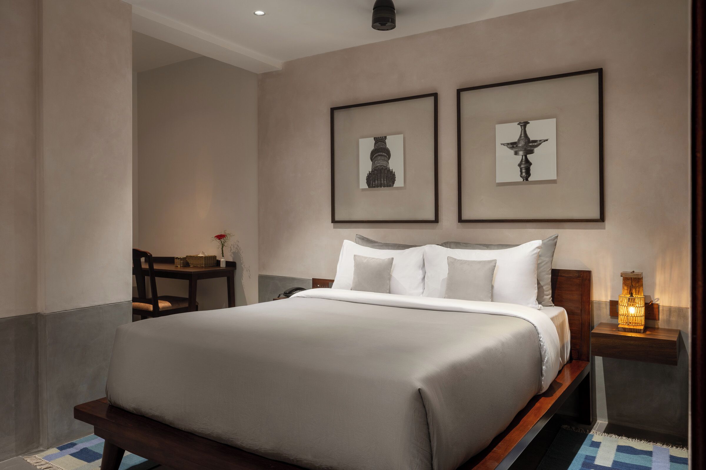 Una cama bien hecha con cabecera de madera, una mesita de noche con lámpara y dos cuadros enmarcados en la pared.