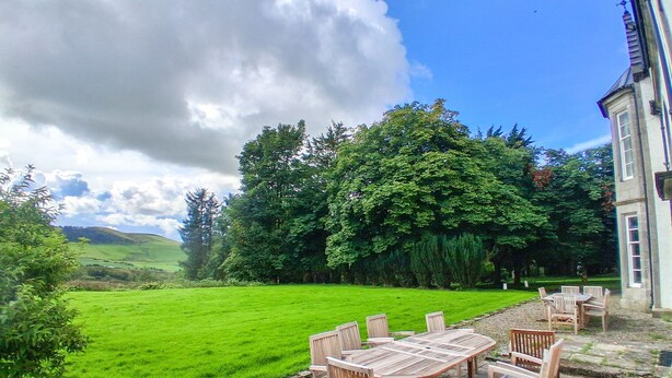 A patio with a wooden table and chairs, surrounded by a lush green lawn and trees, with a building in the background. -  Hillside House , 18th century secluded country house with hot tub & gardens. (Dunfermline)