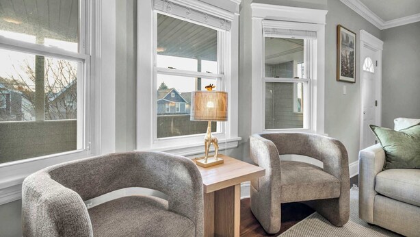 Ein Zimmer mit großen Fenstern, einem Holztisch, zwei Sesseln, einem Sofa und einer Lampe.
