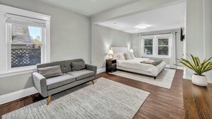 Ein Schlafzimmer mit einem Bett, einem grauen Sofa, einem Nachttisch mit Lampe und einem Fenster mit Jalousien.