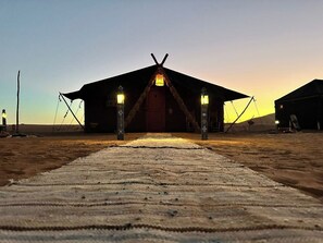 Estrutura tipo tenda com porta central e duas lanternas laterais, situada no deserto, com um caminho revestido de carpete que a conduz.