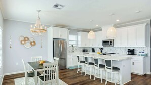 A modern kitchen with white cabinetry, a central island with bar stools, a chandelier, and a stainless steel refrigerator. - Lake2Sea - Spacious Retreat, Gulf & Waterway View! (Surfside Beach)