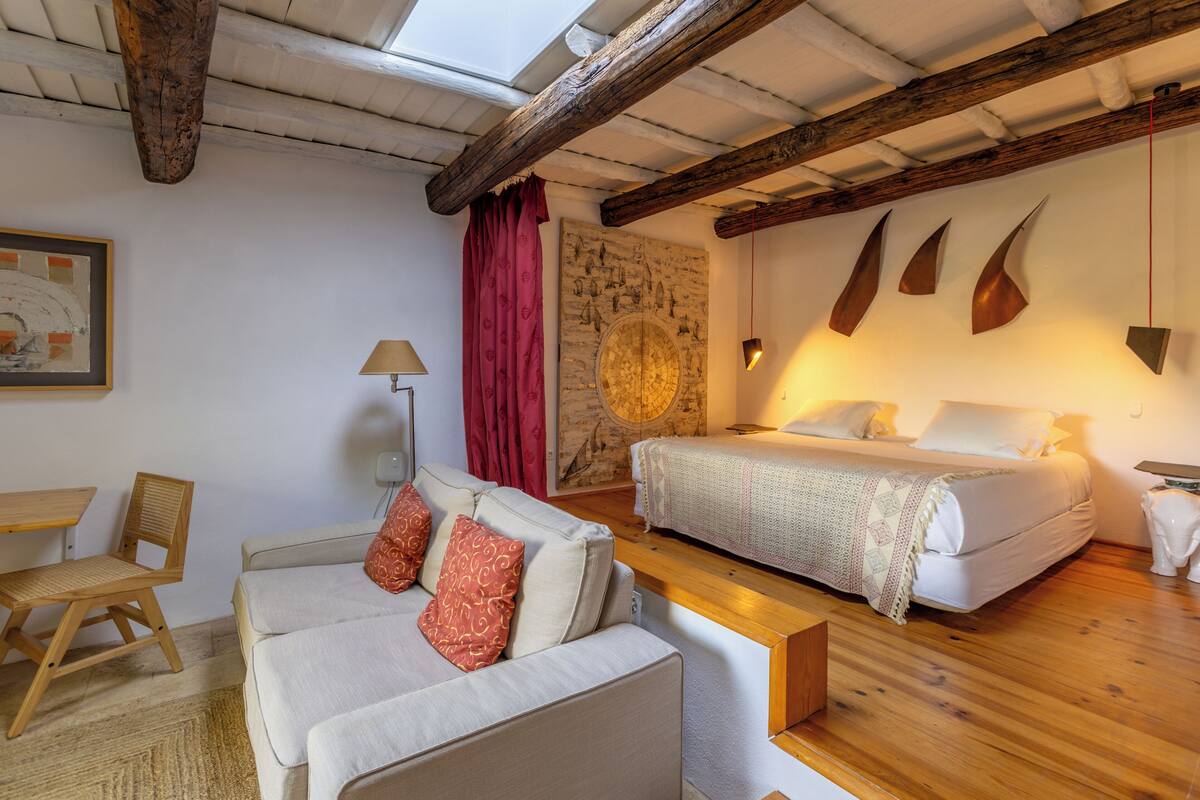 A bedroom with a bed, a sofa, a chair, and a wooden floor.