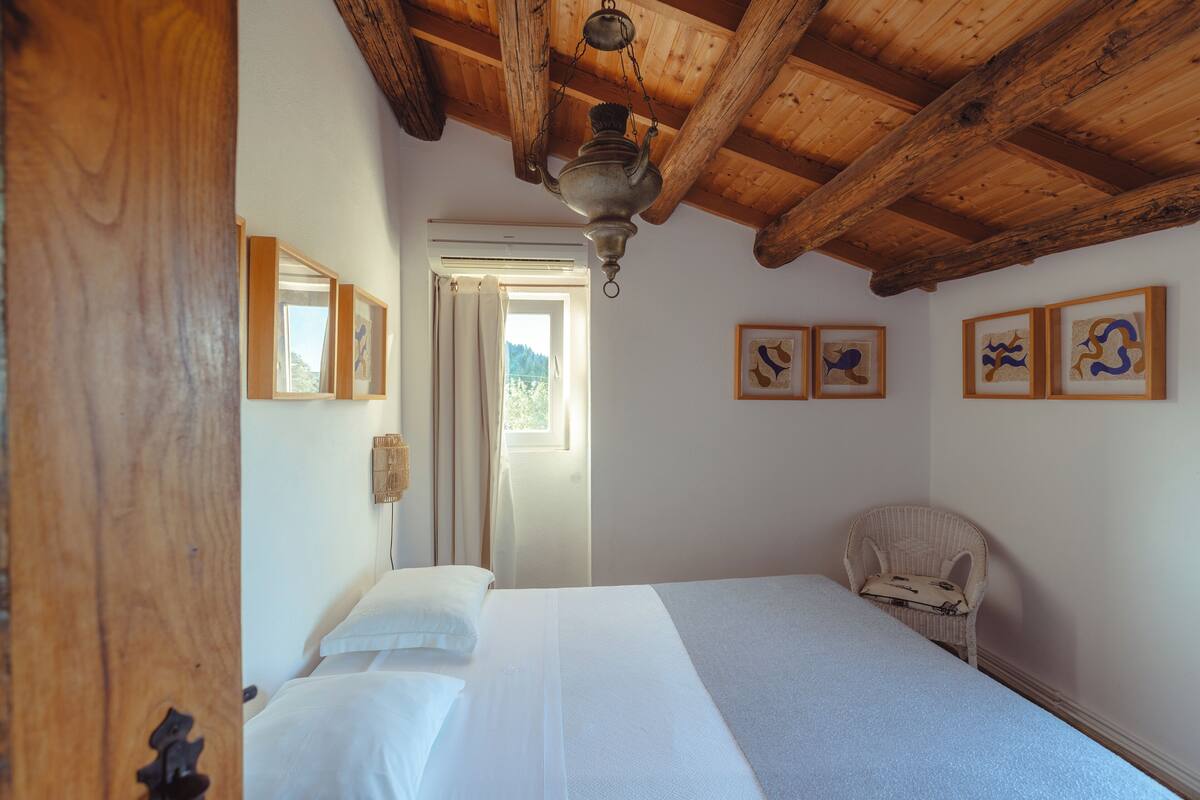 A bedroom with a wooden ceiling, a bed with white bedding, a wicker chair, and framed artwork on the wall.