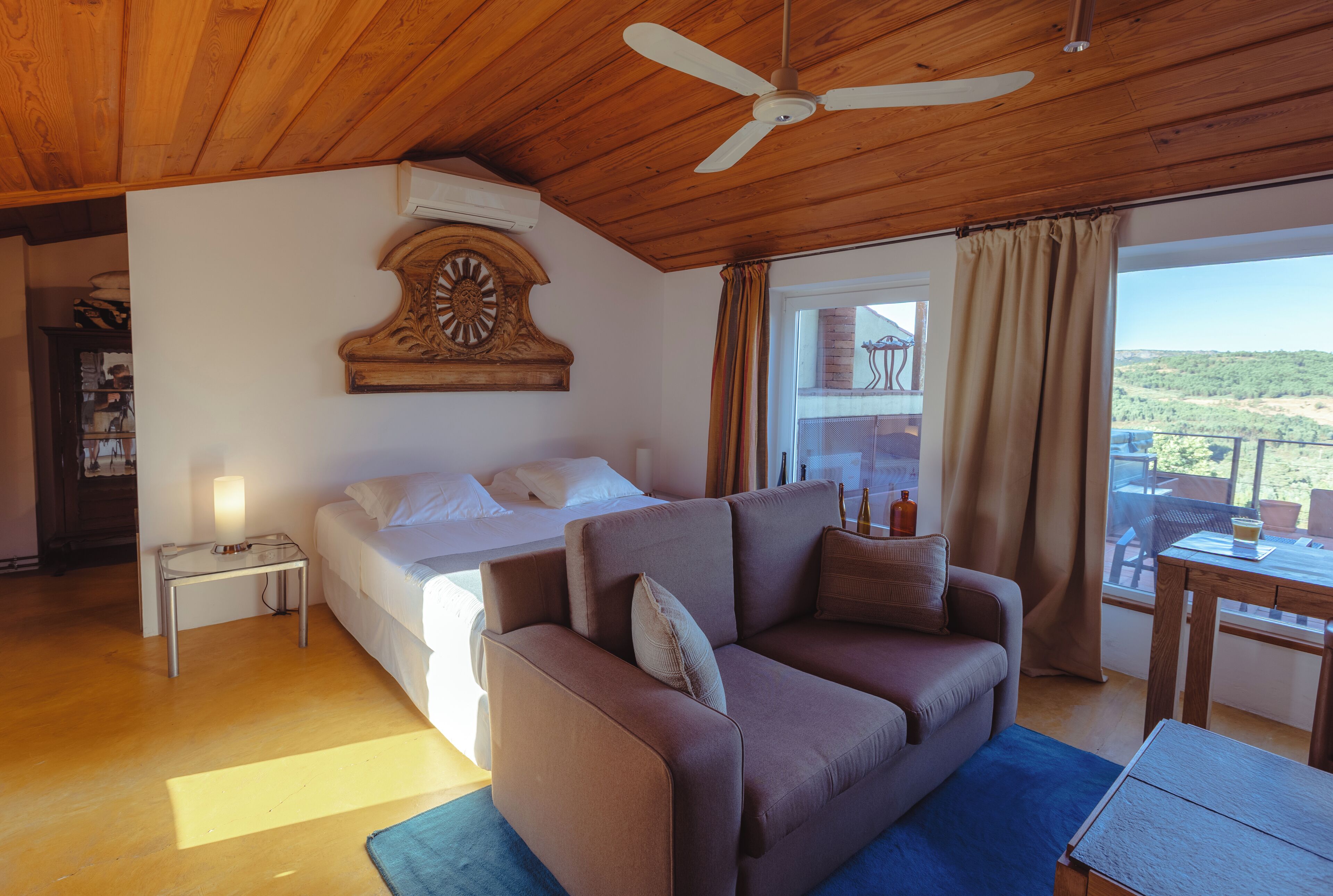 Ein Schlafzimmer mit Bett, einer Couch, einem hölzernen Deckenventilator, einer großen Uhr und Blick ins Freie.