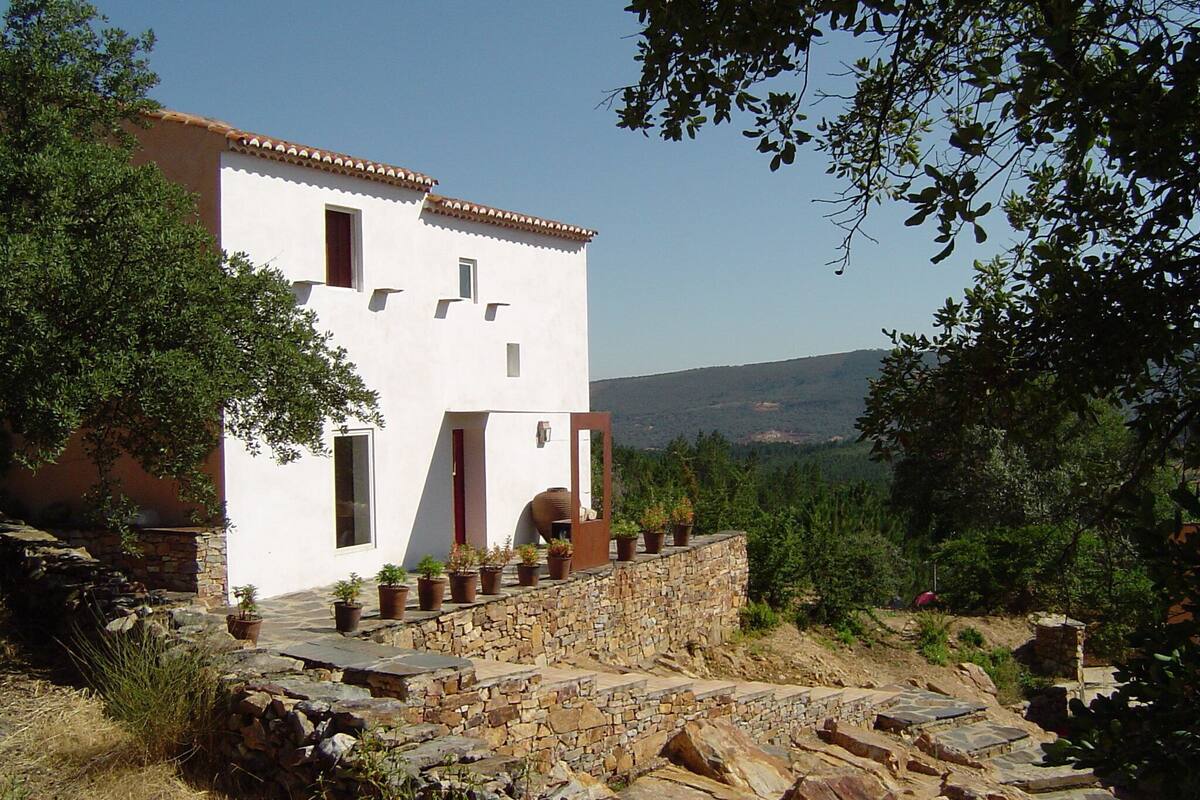 A white building with a stone staircase leading up to a door, surrounded by greenery and a view of hills.