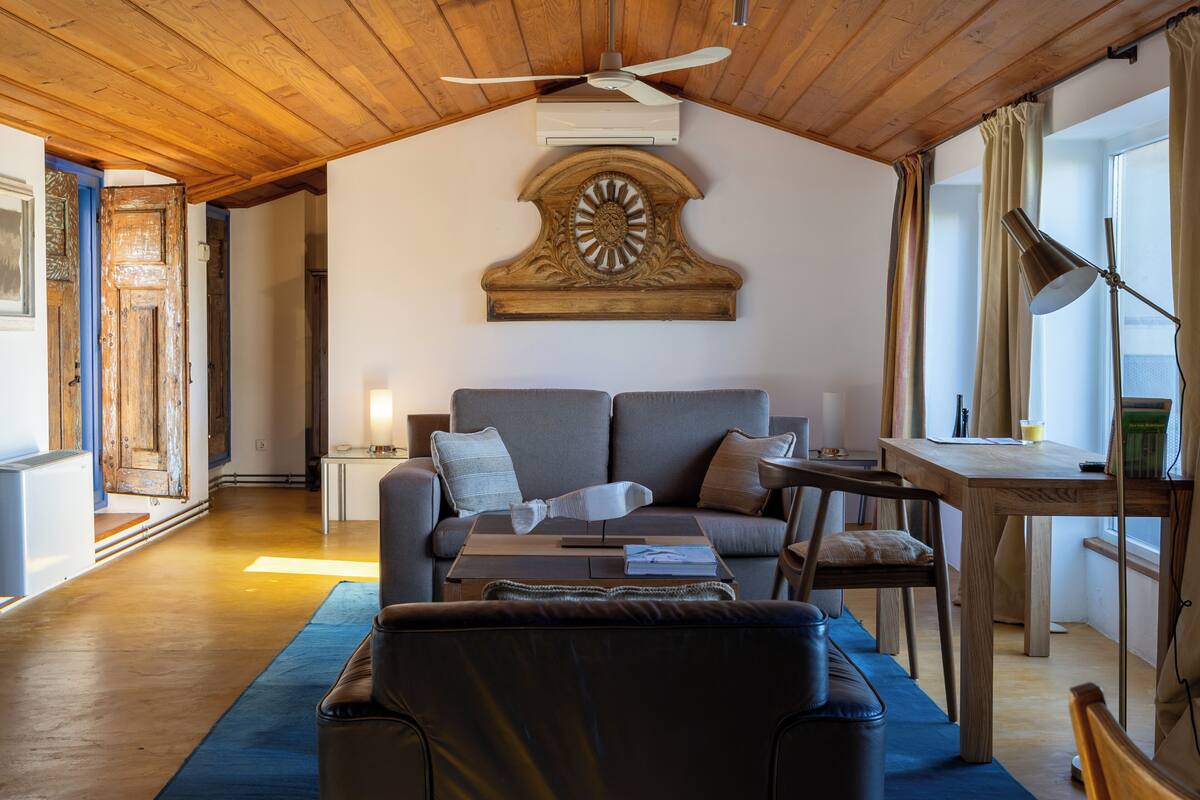 A cozy living room with a wooden ceiling, a large wall clock, a sofa, a coffee table, and a desk with a lamp.