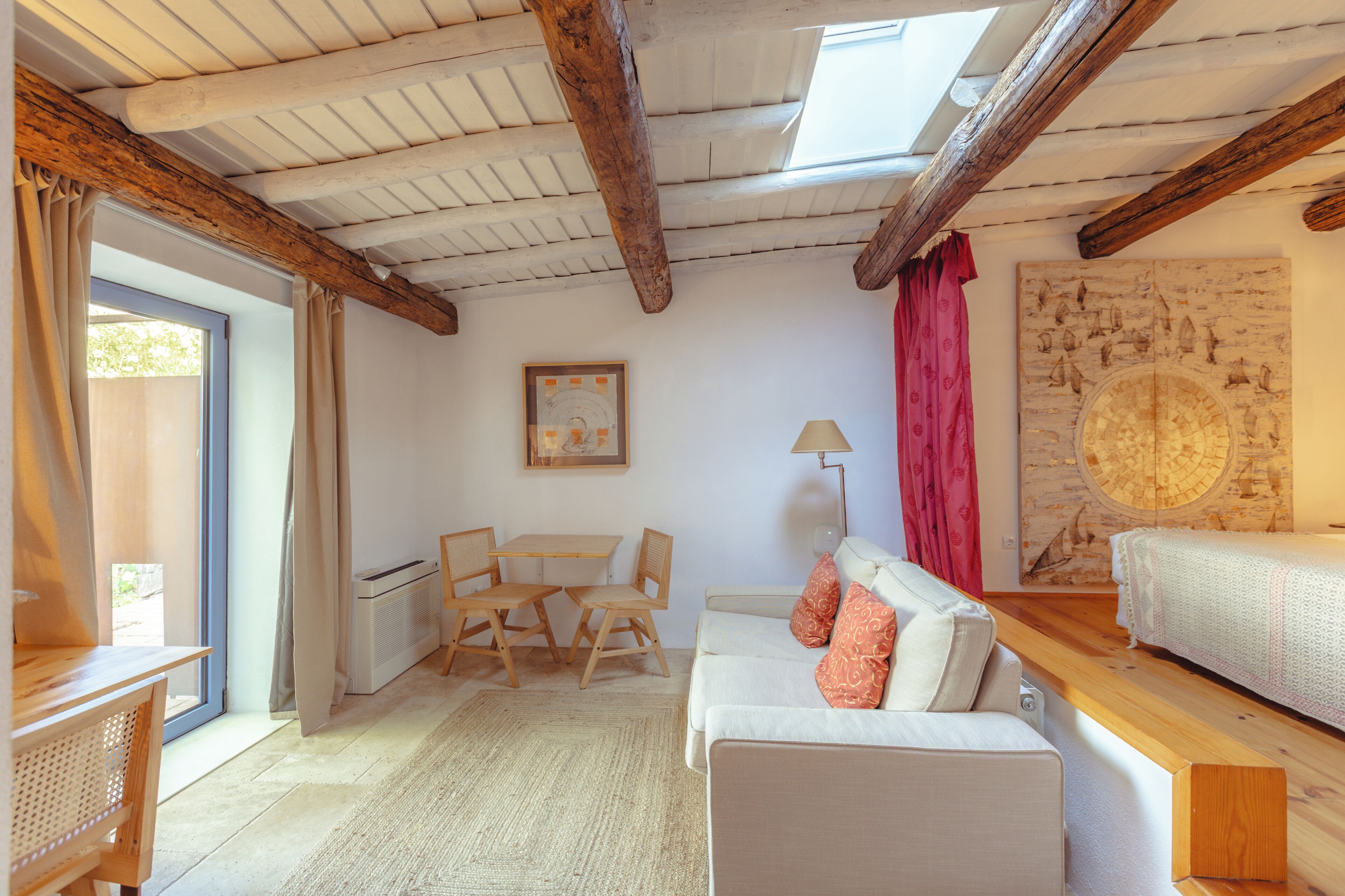 A cozy room with a wooden ceiling, a white sofa with orange pillows, a dining table with chairs, and a large window with curtains.