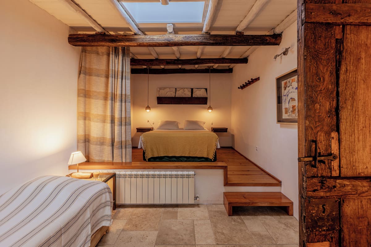A bedroom with a skylight, a bed with a striped bedspread, a bedside table with a lamp, a radiator, and a wooden staircase.