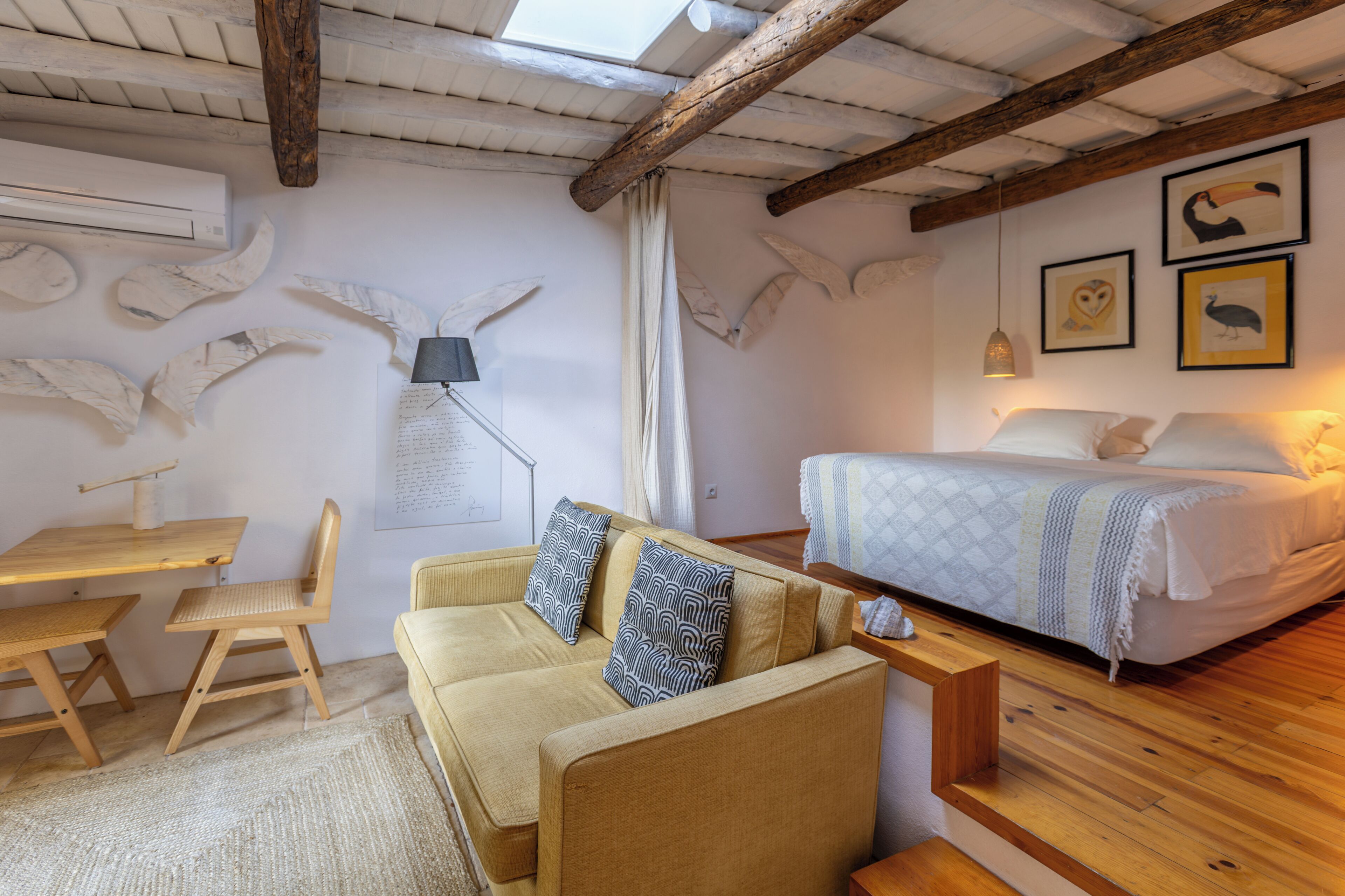 Una habitación acogedora con techo de madera, una cama con ropa de cama blanca, un sofá con cojines estampados, una mesa de madera con sillas y decoración en las paredes.