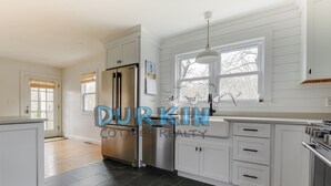 Cocina moderna con armarios blancos, refrigerador de acero inoxidable, lámpara colgante y ventana con vista a árboles.