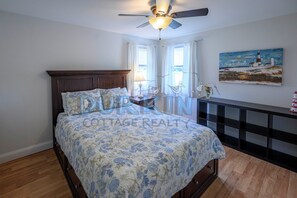 Un dormitorio con cama, ventilador de techo, un cuadro y una ventana con cortinas.