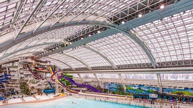 Ein großer Indoor-Wasserpark mit einem Schwimmbecken, mehreren Wasserrutschen und einer hohen, gläsernen Deckenkonstruktion.