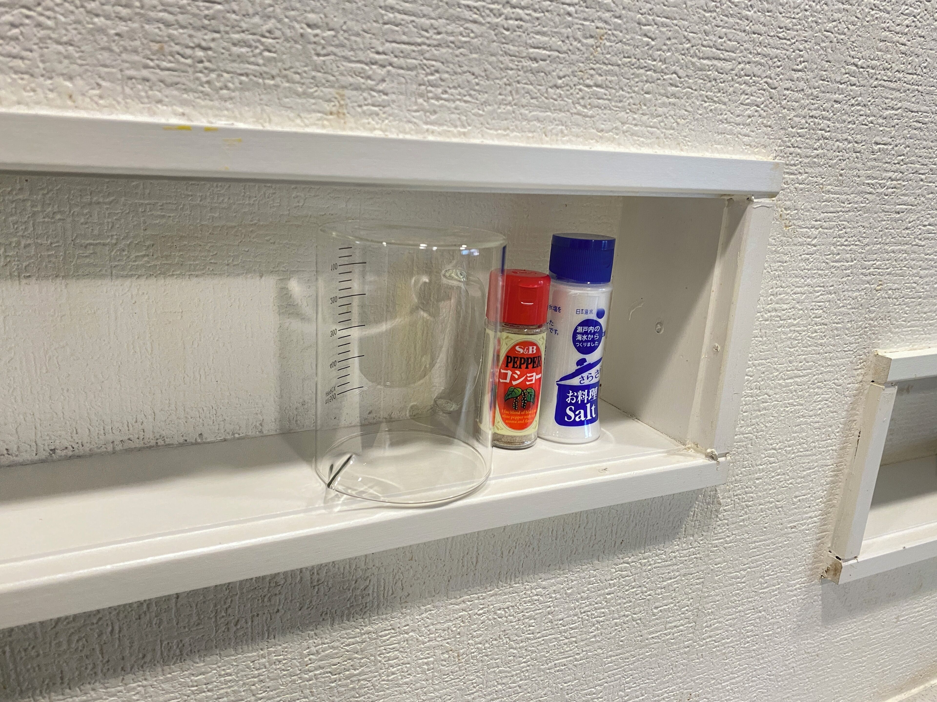 A shelf with a measuring cup, a bottle of S&B Pepper, and a bottle of salt.