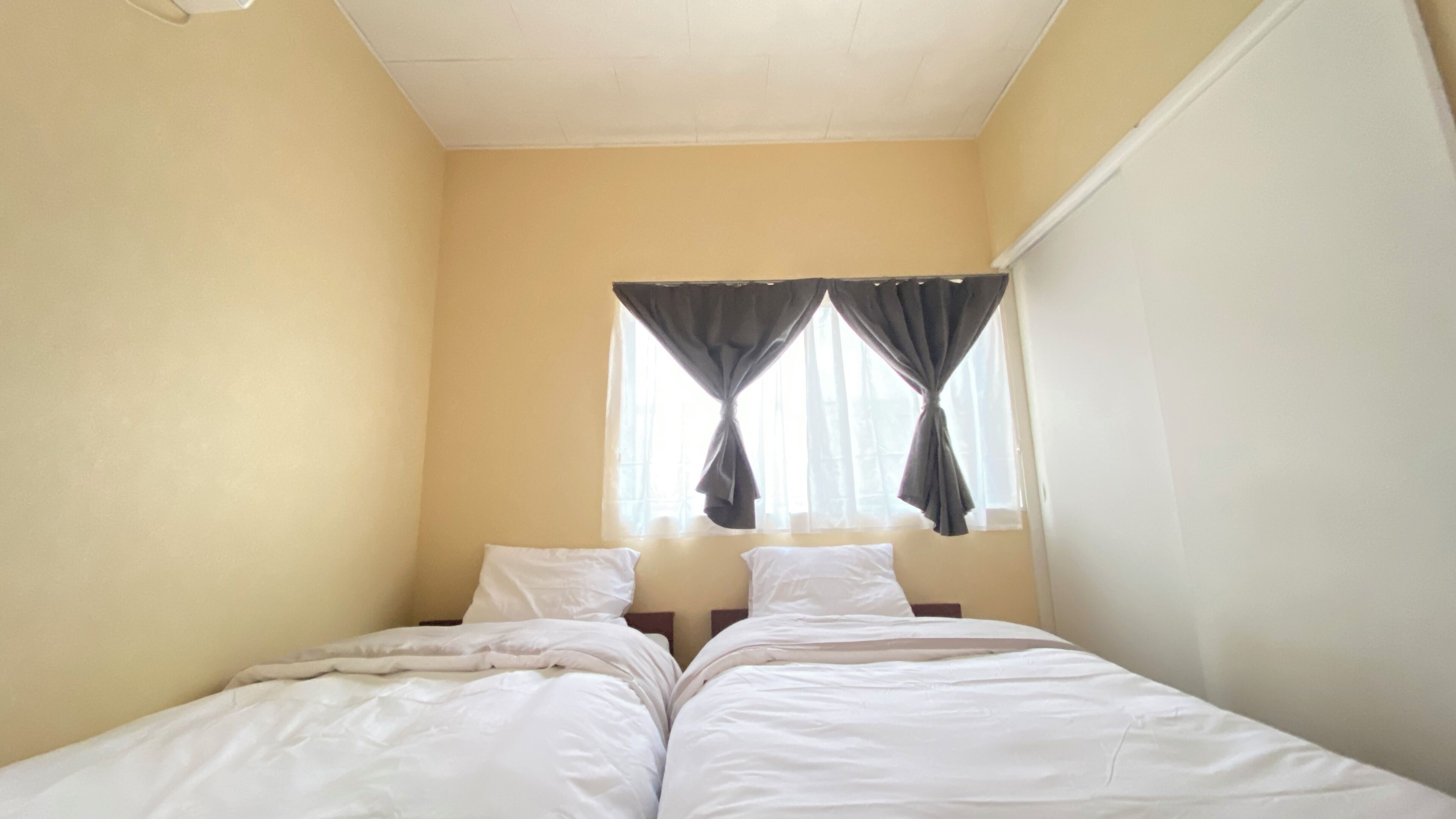 Een kleine kamer met een enkel bed, een effen gele muur, een raam met gordijnen en een wit plafond.